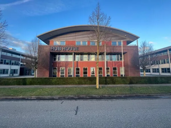 Voorzijde van het moderne kantoorgebouw "HUB HOUSE" met roodbruine gevel en omliggend groen.